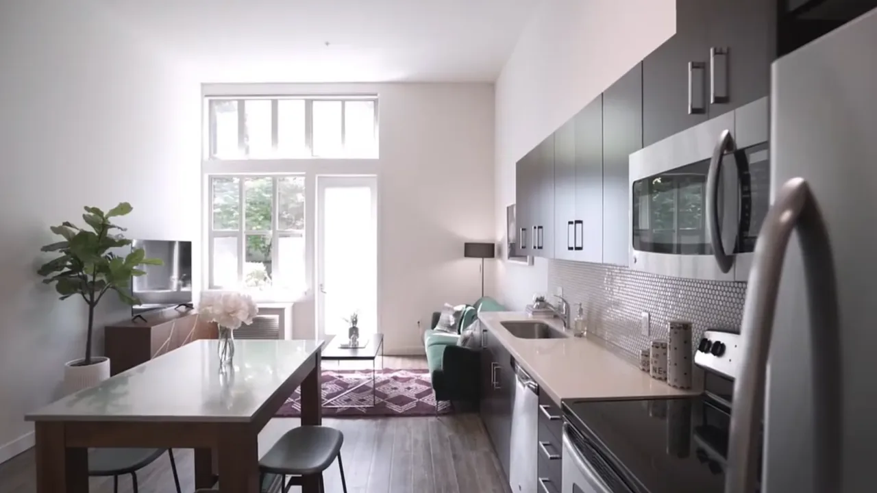 Modern kitchen-living area with sleek cabinets, dining table, and cozy seating. Large windows illuminate room, creating a bright and inviting atmosphere.